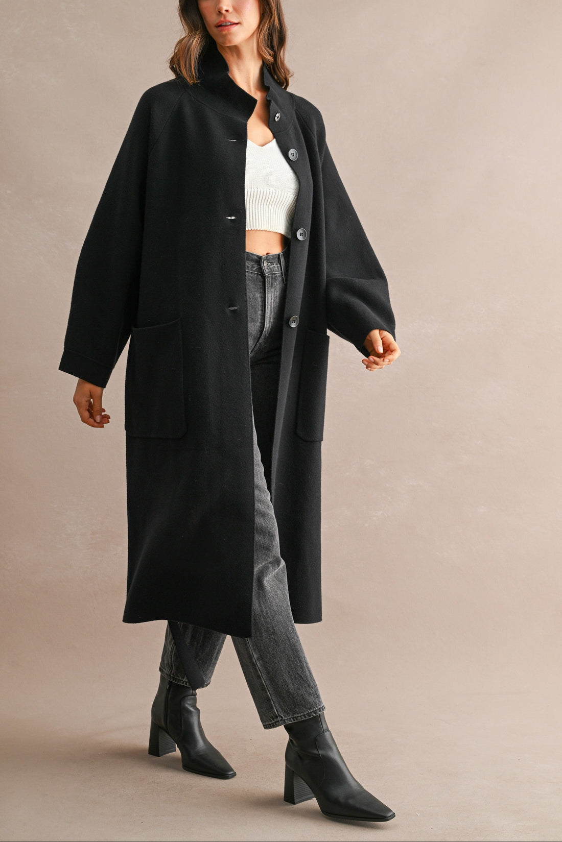 Model wearing black longline coat open over a white knit top, dark jeans, and ankle boots