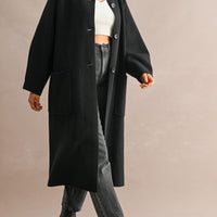 Model wearing black longline coat open over a white knit top, dark jeans, and ankle boots