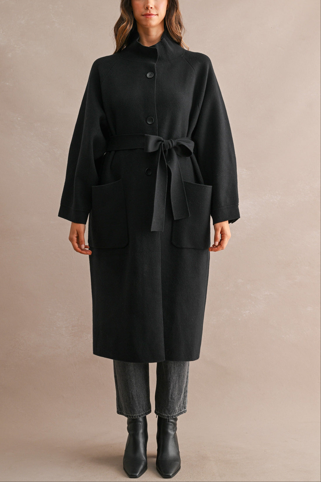 Model wearing black stand collar coat with waist belt tied at the front and hands in patch pockets
