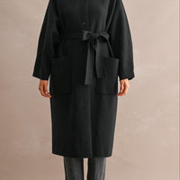Model wearing black stand collar coat with waist belt tied at the front and hands in patch pockets